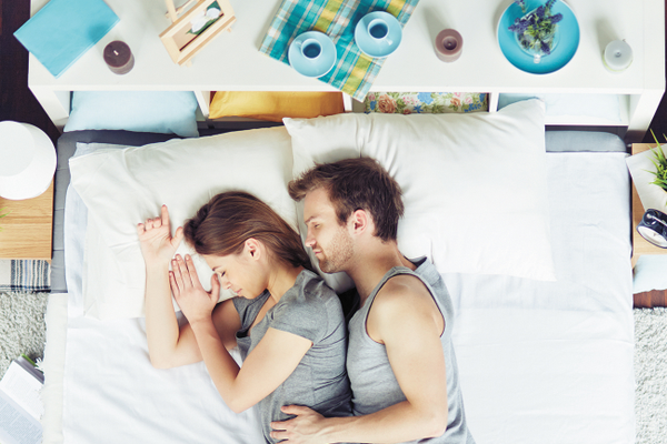 Couple sleeping in bed on their sides embracing on white sheets with pillows, candles, mugs, and a picture frame above their headboard behind them.