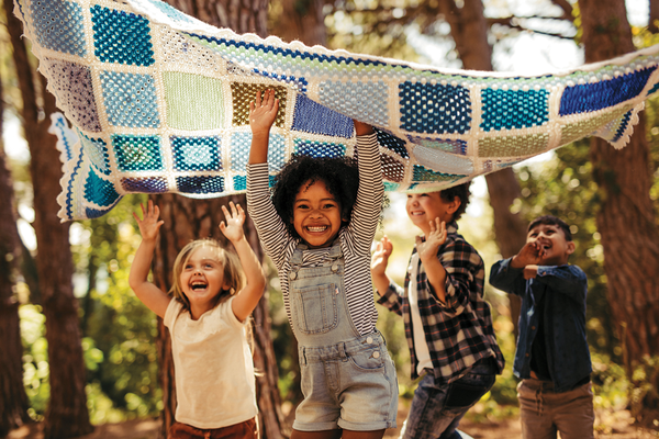 Group of children running in the woods under a quilt with their hands raised laughing and smiling