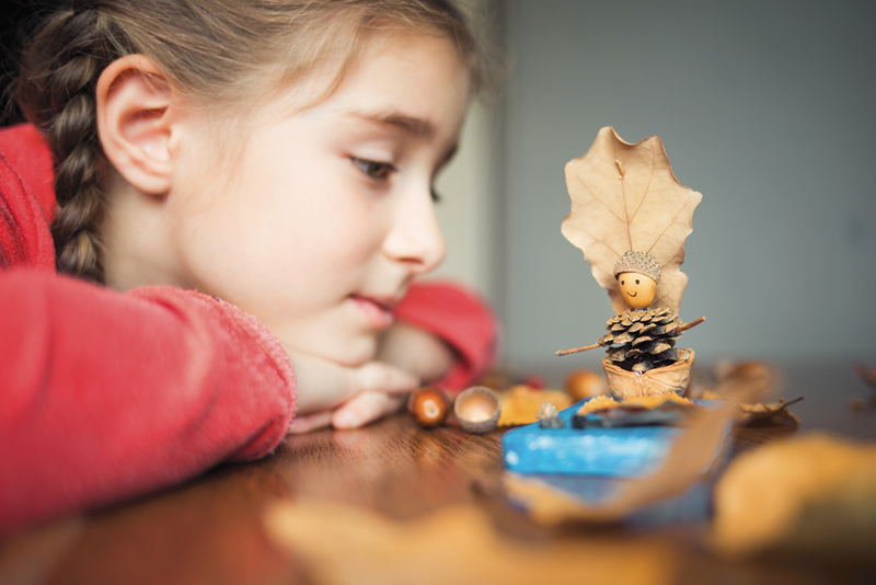 Child leaning on table looking at craft made out of acorns, pinecones, leaves and twigs
