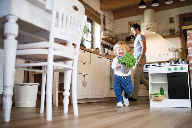Small child carrying potted plant in kitchen with parent in background