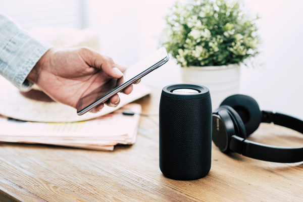 Hand holding out smart phone next to bluetooth speaker and headphones sitting on table next to papers and potted plant