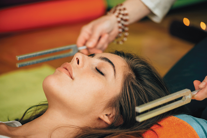 Person lying down with eyes closed while practitioner holds tuning forks alongside head for sound healing